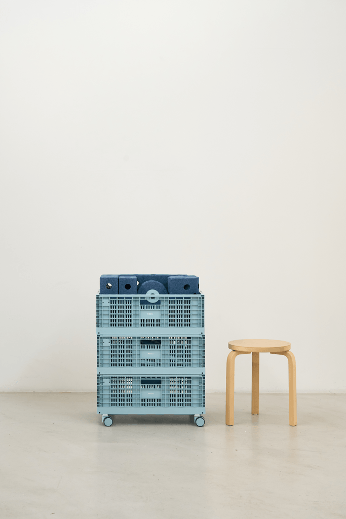 Three blue MODU crates stacked on castor wheels beside a wooden stool, with blue modular blocks in the top crate.