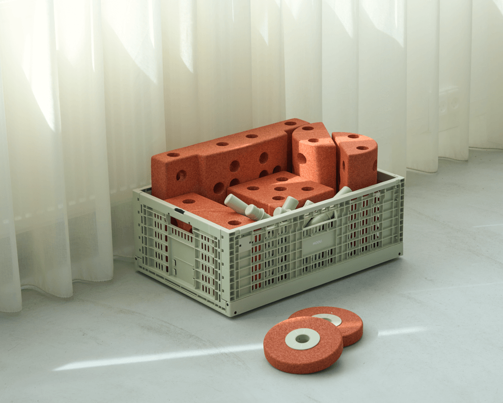 Sage-green MODU crate filled with orange modular blocks beside sheer white curtains, with two orange wheels on the sunlit floor.