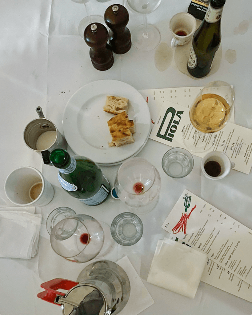 Piola menus among wine glasses, espresso cups, bottles and bread on a white tablecloth marked with spills.