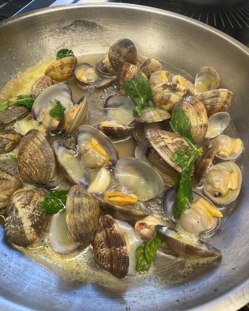 Clams in their shells with green herbs and sauce in a metal cooking pan.