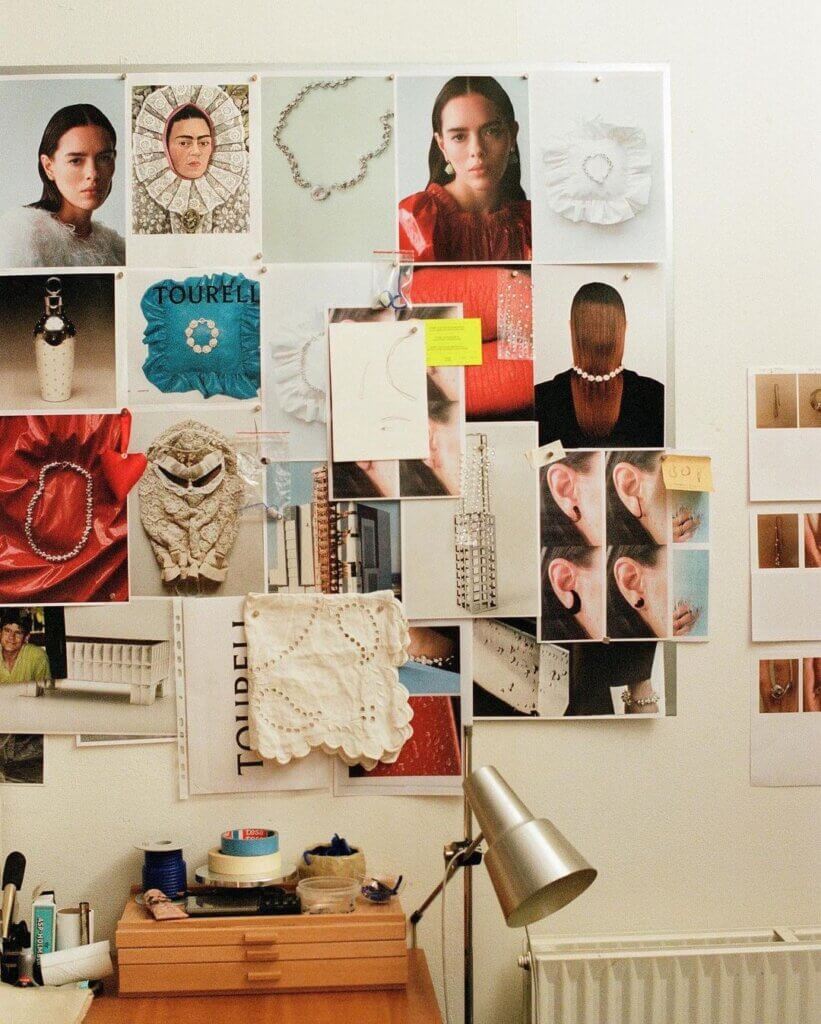 Wall above a desk covered with jewelry photographs, portraits, sketches and a textured fabric swatch.