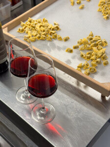 Two glasses of red wine beside a wooden drying tray filled with small pieces of handmade pasta.