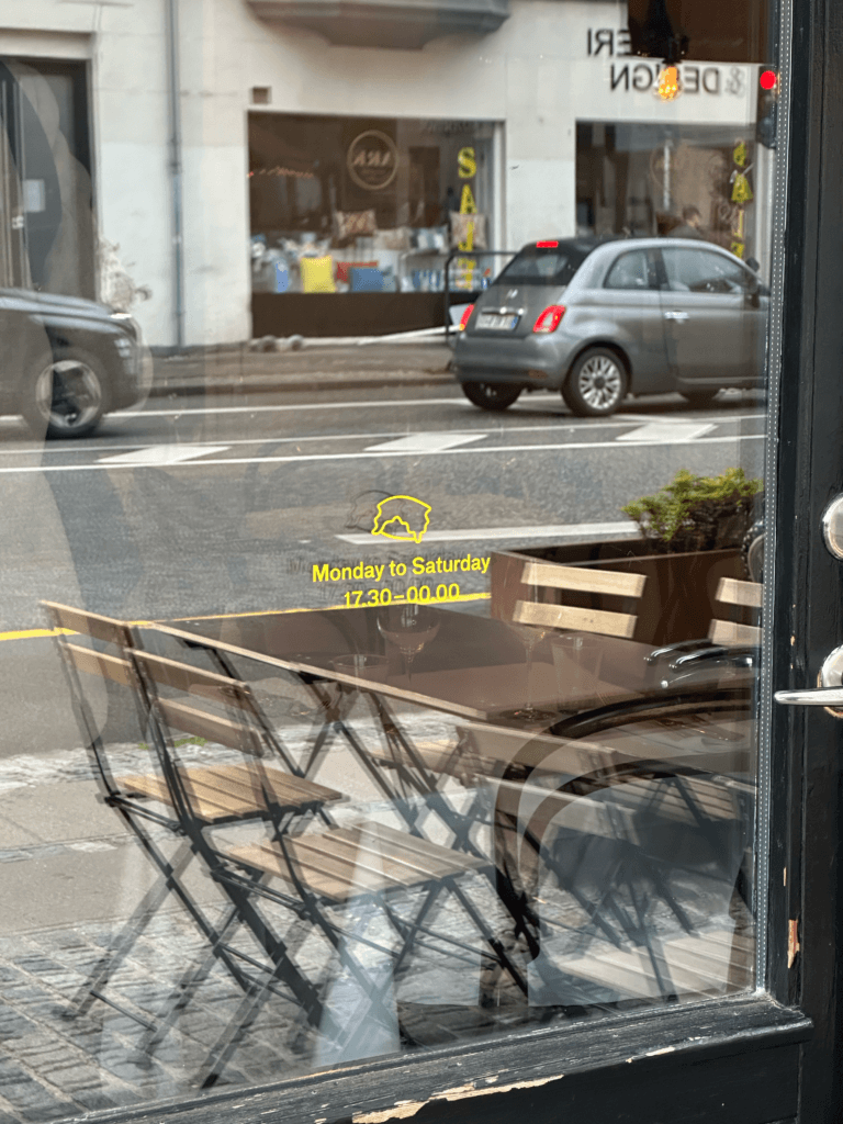 Yellow opening hours and a small outlined symbol on Piola’s glass door, reflecting outdoor tables and the street.