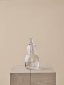Clear glass carafe with an inverted drinking glass on top, standing on a wooden plinth against a beige background.