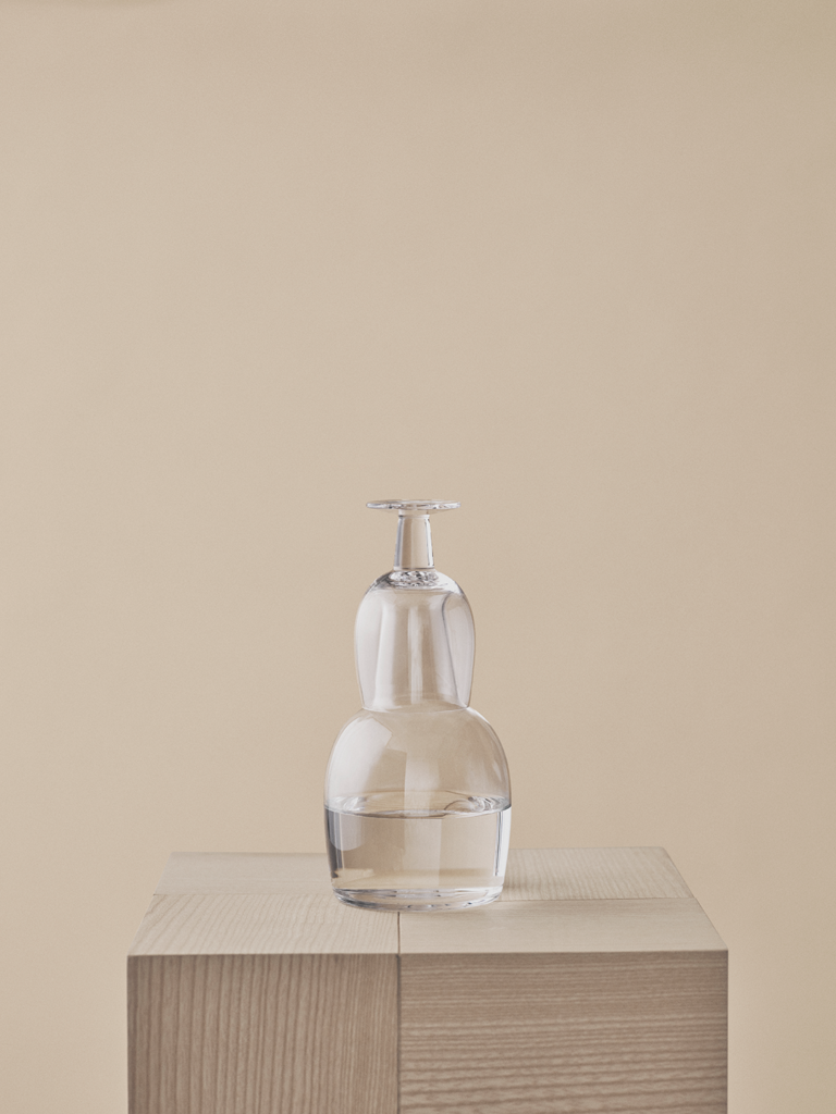 Clear glass carafe with an inverted drinking glass on top, standing on a wooden plinth against a beige background.