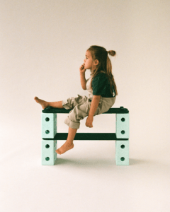 A child sits in profile on a tall MODU structure of green-coloured beams and mint-coloured blocks, one foot dangling, against a cream-coloured backdrop.