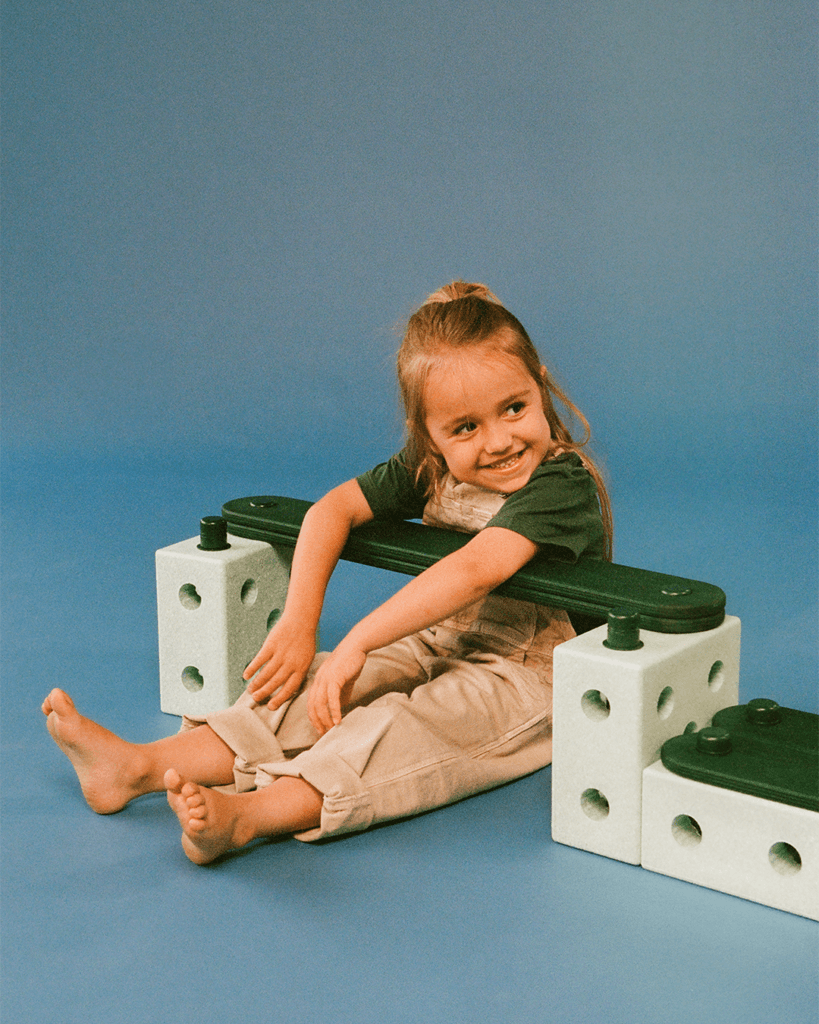 A smiling child in beige dungarees sits behind a green-coloured MODU beam, arms draped over it and legs outstretched against a blue backdrop.