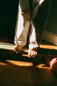 Close-up of bare feet on a green-coloured MODU balance beam, toes extending over the edge, with sunlight and shadows across the parquet floor.