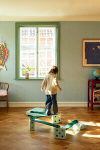 A child faces a window while balancing on green-coloured MODU beams arranged in a zigzag across a sunlit parquet floor.