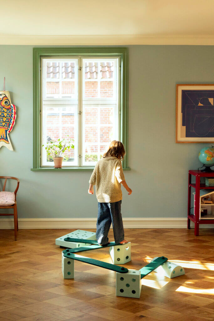 A child faces a window while balancing on green-coloured MODU beams arranged in a zigzag across a sunlit parquet floor.