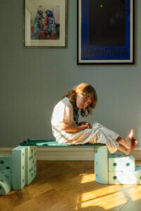 A child sits sideways on a raised green-coloured MODU beam, bare feet outstretched, with sunlight falling across the wooden floor.