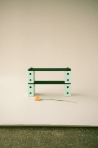 Two green MODU beams stacked on mint-coloured blocks, with an orange flower on a cream paper backdrop whose lower edge reveals the studio floor.