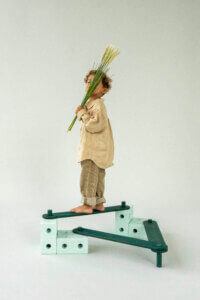 A barefoot child holds tall grasses in front of their face while standing on a green-coloured MODU balance beam against a pale background.