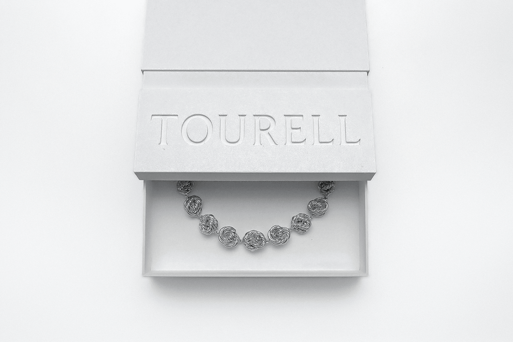 Partly open, textured white Tourell box with recessed lettering on the lid, revealing a necklace of silver-colored, looping forms.