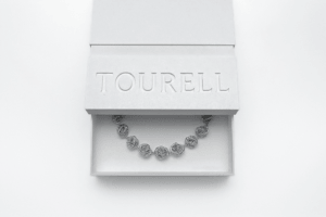 Partly open, textured white Tourell box with recessed lettering on the lid, revealing a necklace of silver-colored, looping forms.